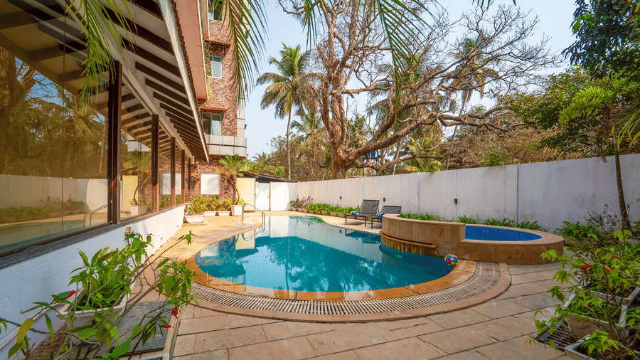 Wide-angle view of Rosetum Anjuna's outdoor pool and jacuzzi surrounded by garden plants, lounge chairs, white boundary wall, and the hotel's distinctive facade.
