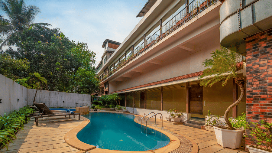 Daytime exterior view of Rosetum Anjuna showing the curved outdoor pool, multi-level hotel balconies, palm trees, and poolside lounge chairs.