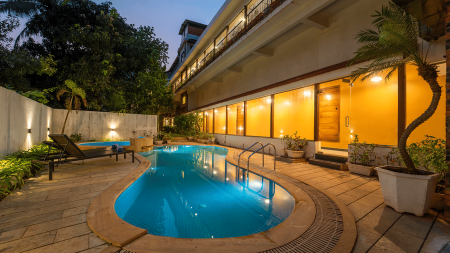 Night view of Rosetum Anjuna's outdoor pool with glowing hotel facade, multi-storey balconies, sun loungers, and tropical trees in the background.