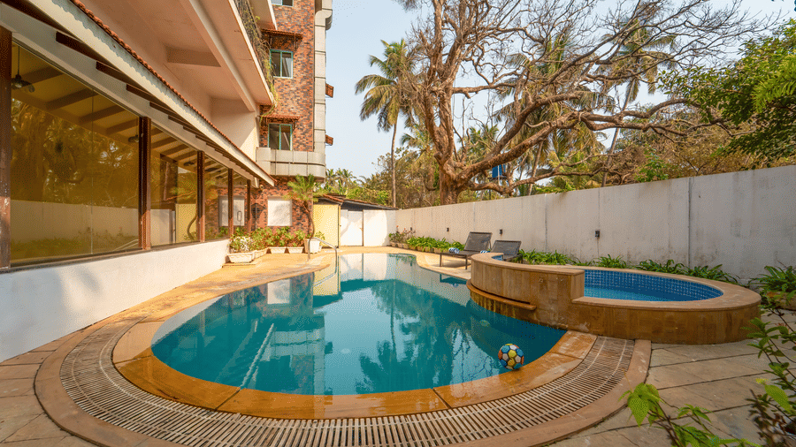 Rosetum Anjuna's kidney-shaped pool and raised circular jacuzzi in daylight, flanked by lounge chairs, a colourful beach ball in the water, and mature trees.
