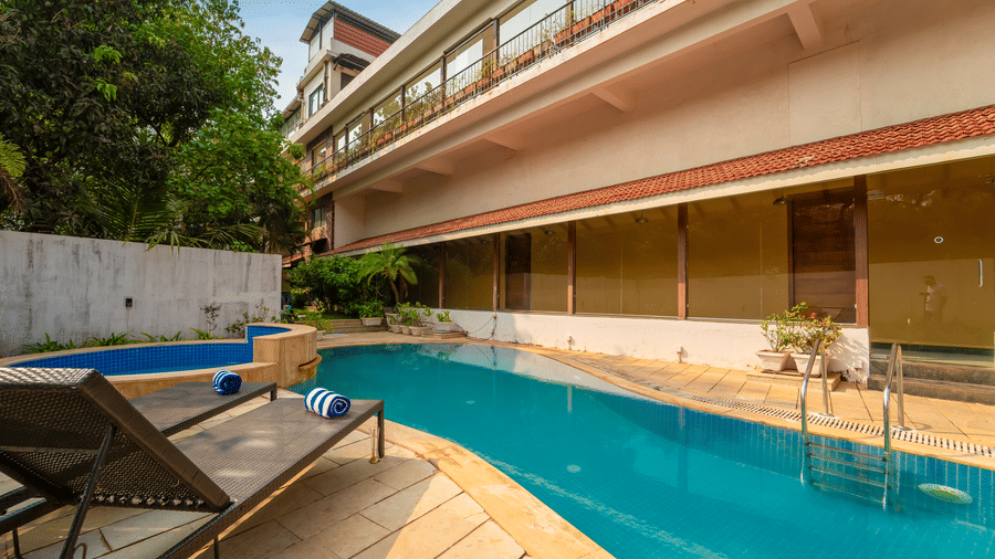 Close-up of Rosetum Anjuna's outdoor pool with two sun loungers and rolled towels in the foreground, a jacuzzi nearby, and the hotel building behind.
