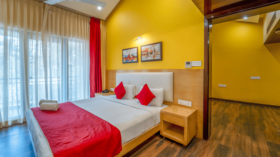 Rosetum Anjuna Royal Suite bedroom with a king bed, red accent pillows, yellow walls, framed artwork, sheer curtains, balcony access, and wooden flooring.