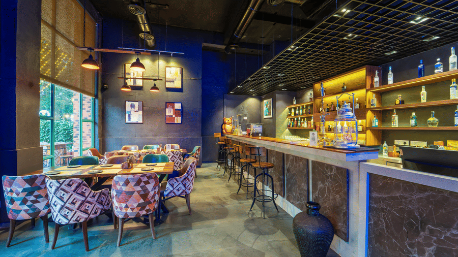 Rosetum Anjuna's Speakeasy Bar showing bar counter with wooden stools, patterned upholstered chairs, backlit bottle shelves, framed wall art, and blue-toned ambience.