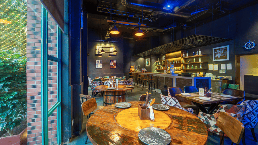  Interior of Rosetum Anjuna's Speakeasy Bar featuring rustic wooden tables, eclectic chairs, a fully stocked back bar, industrial pendant lighting, and blue ambient lighting.