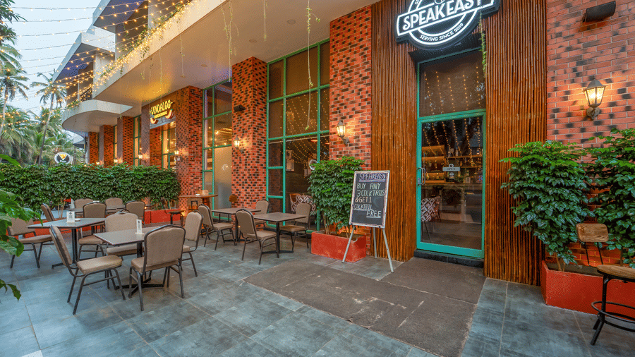 Outdoor entrance of Rosetum Anjuna's Speakeasy Bar with neon signage, fairy lights, al fresco seating, brick facade, bamboo pillars, and a cocktail offer chalkboard.