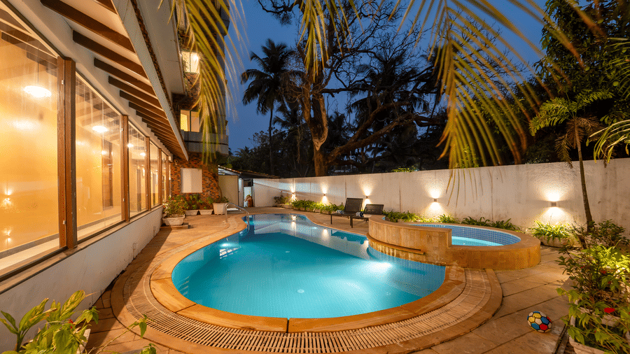 Rosetum Anjuna's kidney-shaped outdoor swimming pool and jacuzzi illuminated at night, surrounded by lush greenery, lounge chairs, and warm wall lighting.