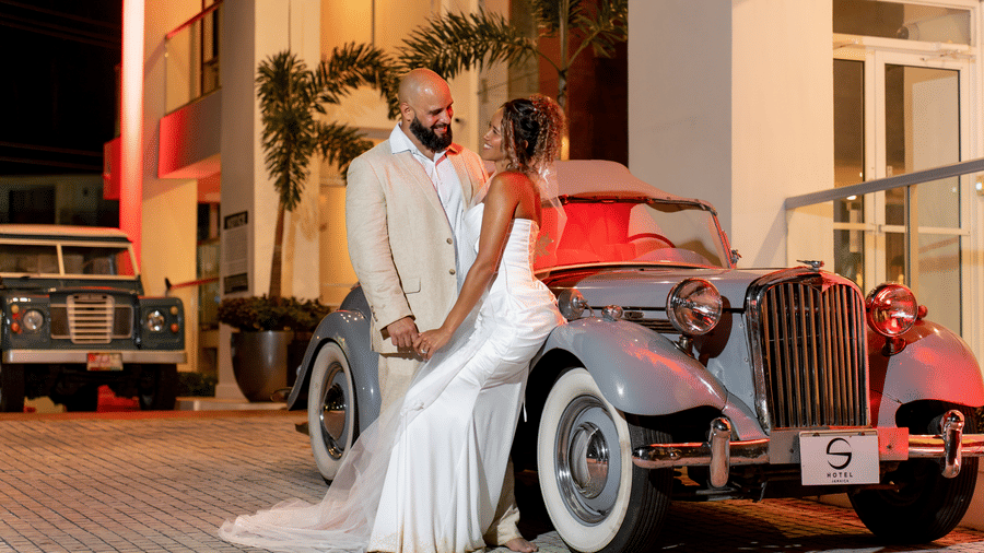 Bride and groom in elegant attire standing beside a vintage car at entrance of S Hotel Montego Bay at night with warm lighting and palm trees creating a romantic setting.
