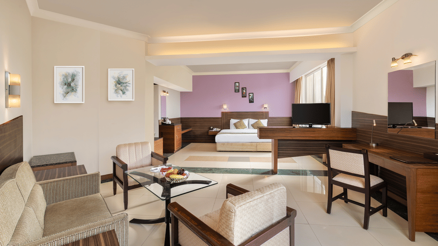 An overview of the Deluxe Rooms in Tanjore at Sangam Hotel, Thanjavur, featuring a double bed in the background with lamps, paintings, and a study table, chairs, and a coffee table in the foreground.