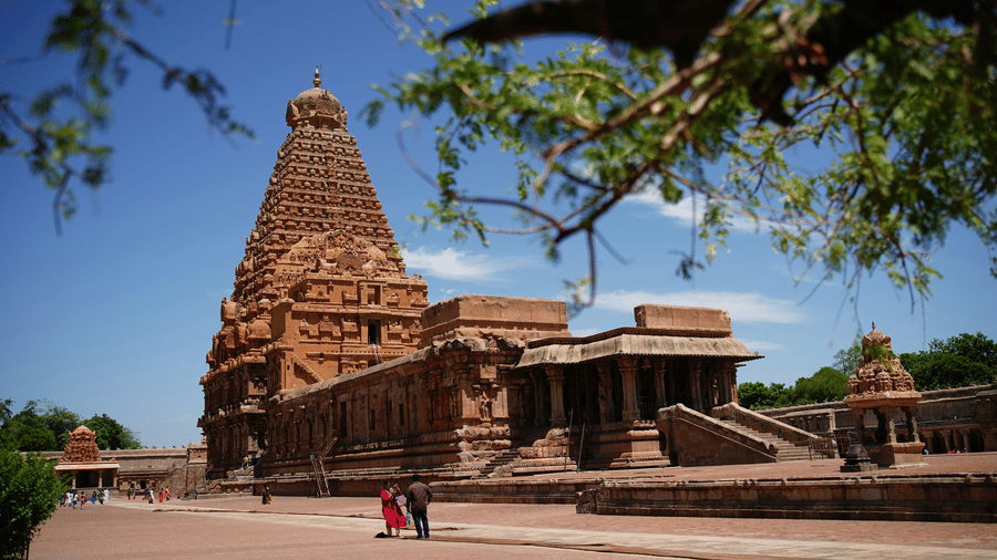 The grand Brihadeeswarar Temple complex stands prominently under a clear blue sky with an open space next to the temple and people walking on it. This temple is one of the popular places to visit during a Thanjavur trip from Chennai.