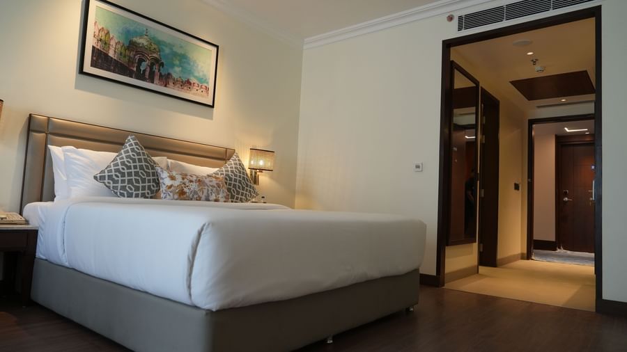 A well-lit hotel room features a bed with white linens and a painting above the headboard, leading to an open doorway on the right | Deluxe Double Bed Room | Sarovar Portico, Hanumangarh
