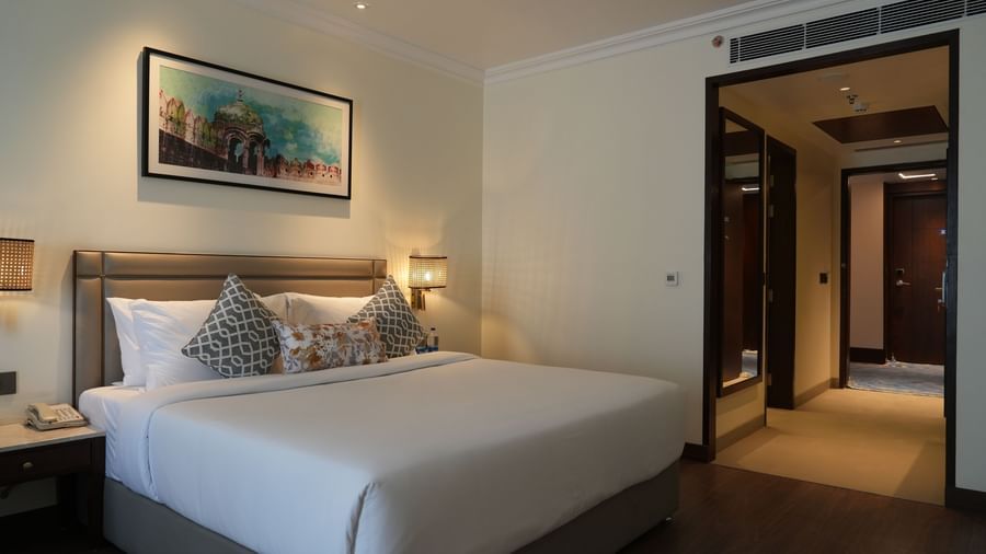 A well-lit hotel room features a bed with white linens and a painting above the headboard, leading to an open doorway on the right | Deluxe Double Bed Room | Sarovar Portico, Hanumangarh