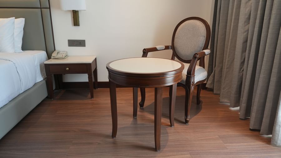 A hotel room with a bed, a bedside table with a lamp, and a chair and small table in the corner | Deluxe Twin Bed Room | Sarovar Portico, Hanumangarh