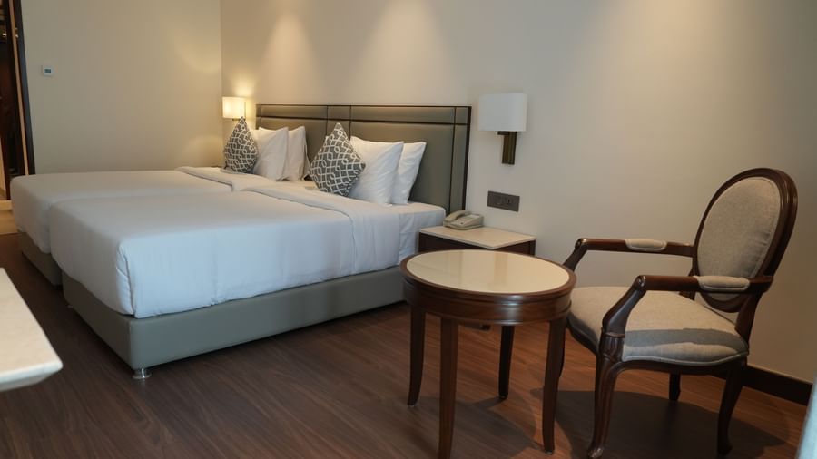 A hotel bedroom highlights a neatly made twin beds, a bedside table with a lamp, and drapes | Deluxe Twin Bed Room | Sarovar Portico, Hanumangarh