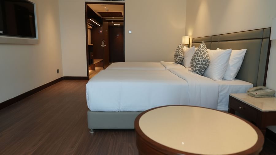 A modern hotel bedroom contains twin beds with white linens, a bedside table, and curtains covering a window | Deluxe Twin Bed Room | Sarovar Portico, Hanumangarh