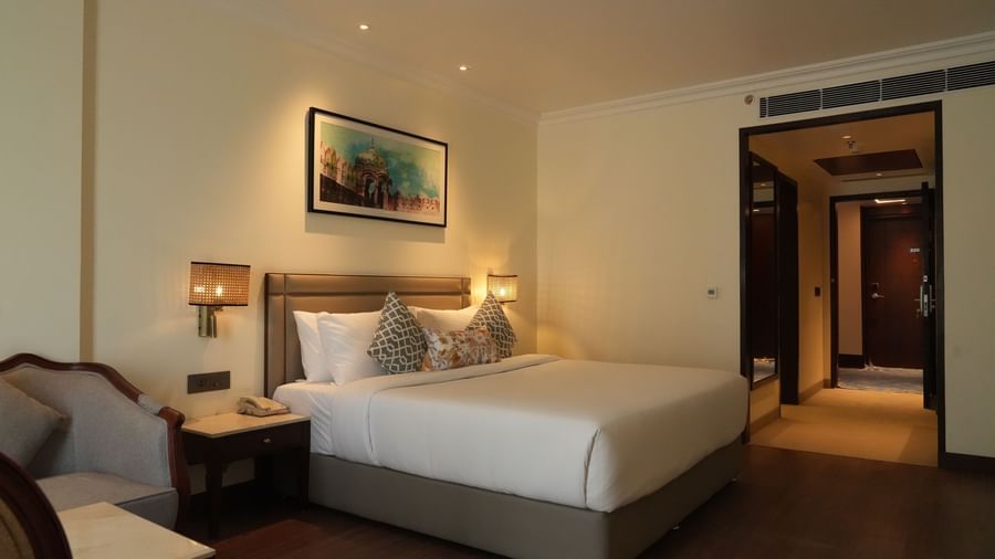 A hotel room with a large bed, a sitting area, and a view into an adjoining space | Deluxe Double Bed Room | Sarovar Portico, Hanumangarh