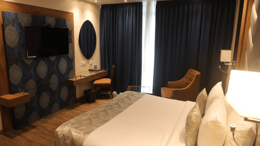Room at Sarovar Portico, Somnath featuring a bed, wall-mounted TV, chairs, and blackout curtains.