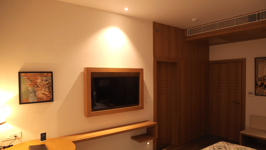 Room corner at Sarovar Portico, Somnath showing a wall-mounted TV, desk, lighting, and wooden wardrobe.