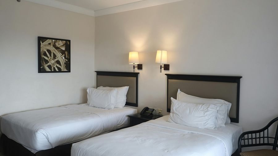 A bright hotel room contains two single beds with white bedding and dark headboards, illuminated by wall-mounted lamps and natural light, with a framed picture on the wall.