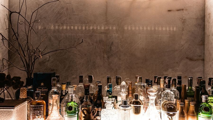 A dimly lit bar shows a counter with illuminated bottles and glasses, set against a textured wall with a faint inscription and bare branches.