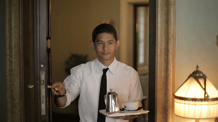 A staff member wearing a uniform and tie holds a tray with a teapot and cup while standing by a doorway next to a lamp.