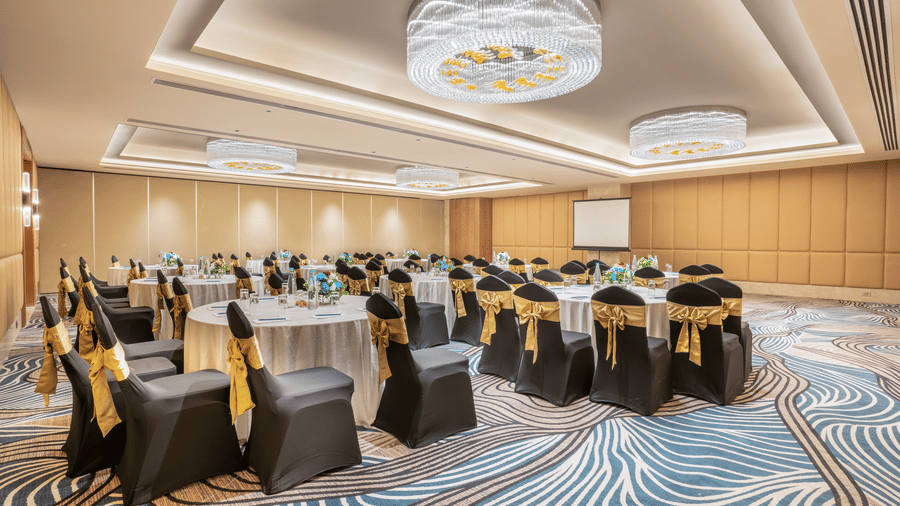 A conference room at Sarovar Premiere Alwar with round tables and chairs with gold sashes and a projector screen.