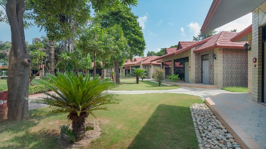 An outdoor image of a property with trees and lawn on one side and the other side has  many rooms adjoined -  Shri Radha Brij Vasundhara Resort & Spa, Mathura