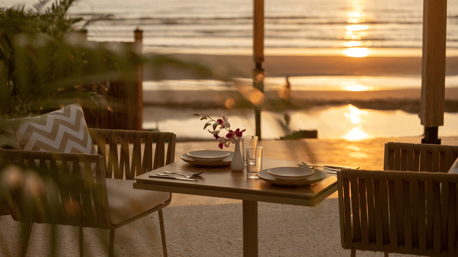 A close up shot of a table with two chairs at saz on the beach restaurant during sunset with sun rays reflecting on the sea waves in the background