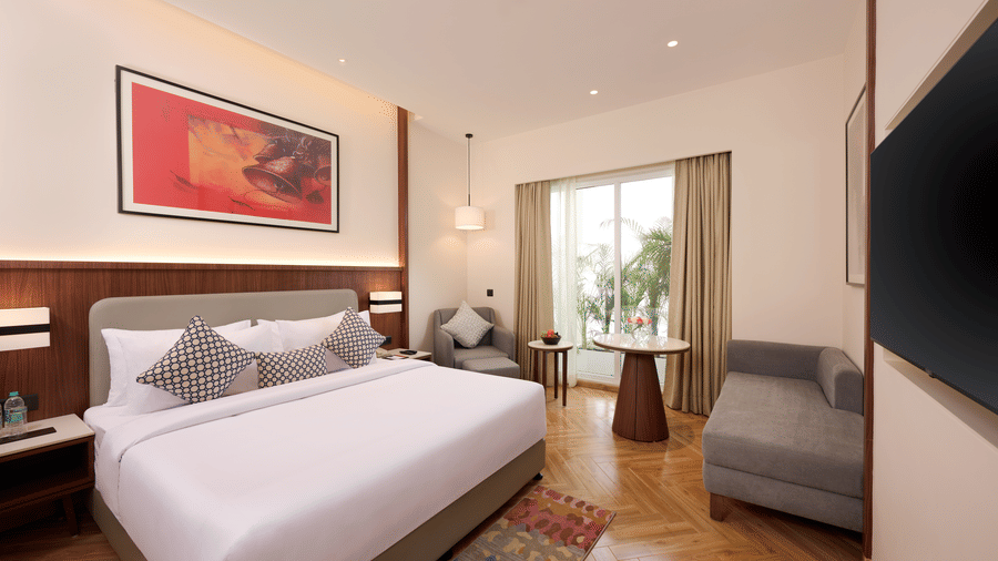 Skywings Sarovar Portico Mathura deluxe king bed with king bed, patterned cushions, large window with natural light, gray sofa, and warm wooden flooring.