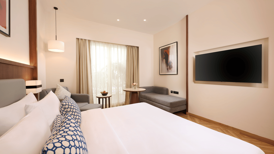 Skywings Sarovar Portico Mathura deluxe king  room wide view with two beds, wall-mounted TV, wooden flooring, large windows, and soft warm lighting.