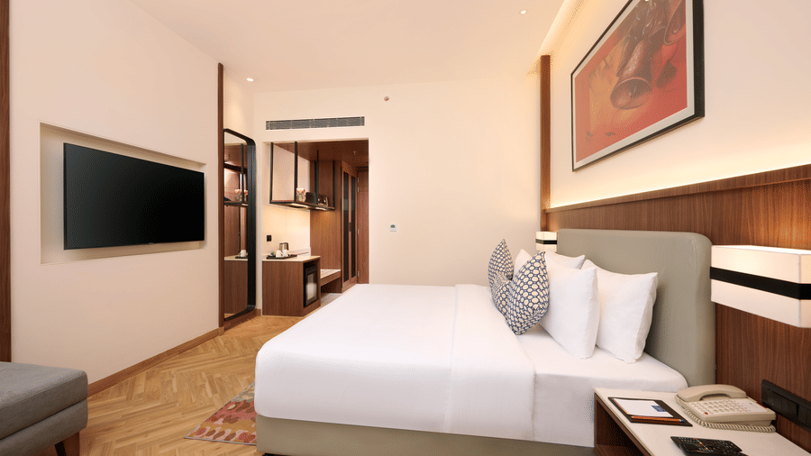 Skywings Sarovar Portico Mathura deluxe bed room with wall-mounted TV, wooden wardrobe, gray sofa, red framed wall art, and herringbone wood flooring.