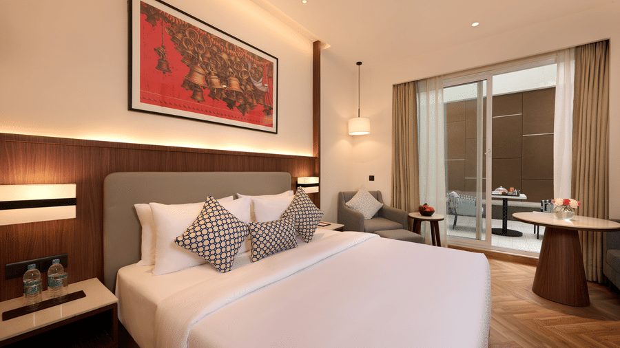 Skywings Sarovar Portico Mathura premium room side view with king bed, patterned cushions, wooden headboard, herringbone flooring, and warm ambient lighting.