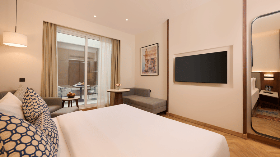 Skywings Sarovar Portico Mathura premium room with king bed, wall-mounted TV, patterned cushions, gray sofa, and bright natural window light.
