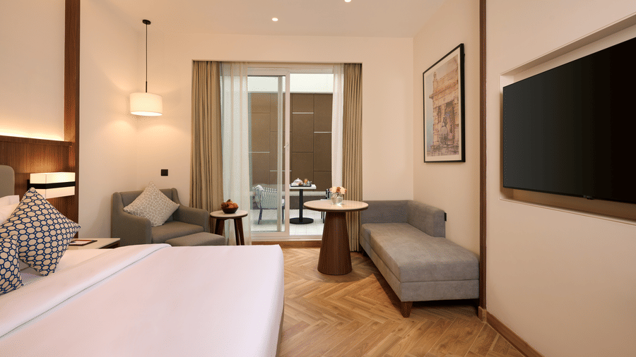 Skywings Sarovar Portico Mathura premium room with gray sofa, wall-mounted TV, wooden accent wall, herringbone flooring, and large draped windows.