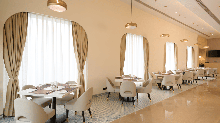 Skywings Sarovar Portico Mathura restaurant with arched draped windows, cushioned dining chairs, round tables, and soft warm natural lighting.
