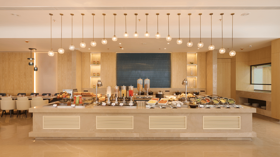 Skywings Sarovar Portico Mathura restaurant with wide buffet counter fully stocked, pendant lighting, blue wall art, and bright cream interiors.