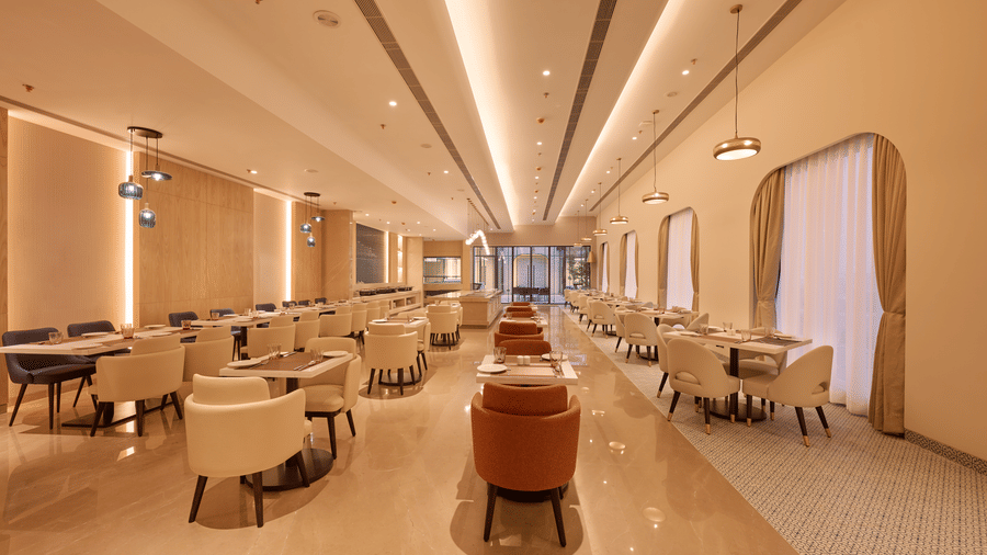 Skywings Sarovar Portico Mathura restaurant corridor with orange and beige chairs, wooden tables, warm recessed lighting, and elegant cream interiors.