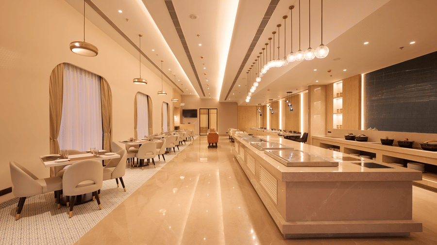 Skywings Sarovar Portico Mathura restaurant central buffet counter at night with pendant lighting, marble flooring, dark display panel, and beige interiors.