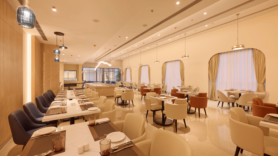 Skywings Sarovar Portico Mathura restaurant with dark chairs along large draped windows, pendant lighting, marble flooring, and warm cream interiors.
