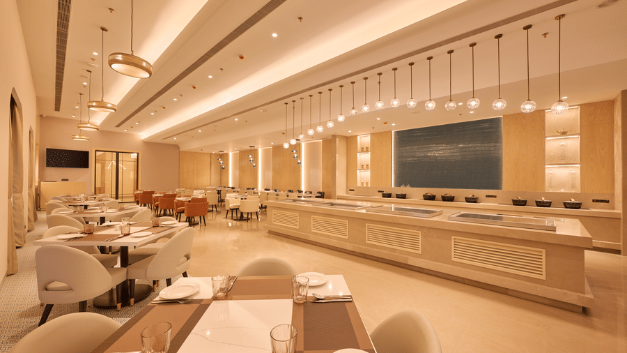 Skywings Sarovar Portico Mathura restaurant dining area with wall-mounted TV, round cushioned chairs, pendant lights, and warm cream-toned interiors.