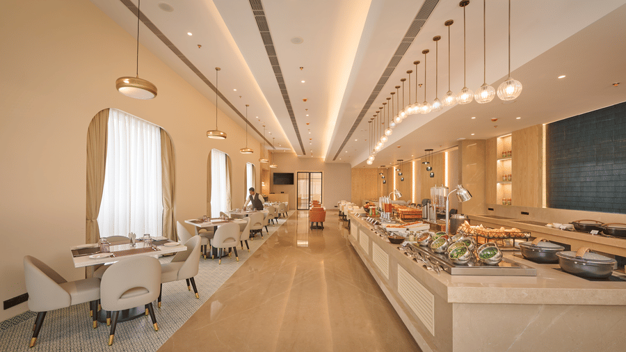 Skywings Sarovar Portico Mathura restaurant empty aisle with pendant lights, marble buffet counter, beige walls, large draped windows, and tiled flooring.