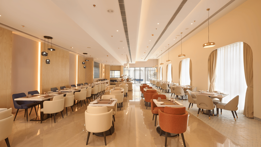 Skywings Sarovar Portico Mathura restaurant in daylight with mixed orange and cream chairs, arched windows, marble flooring, and bright airy ambience.