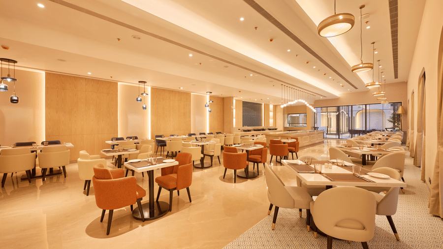 Skywings Sarovar Portico Mathura restaurant with orange and beige cushioned chairs, wooden tables, warm recessed lighting, and cream-toned walls.