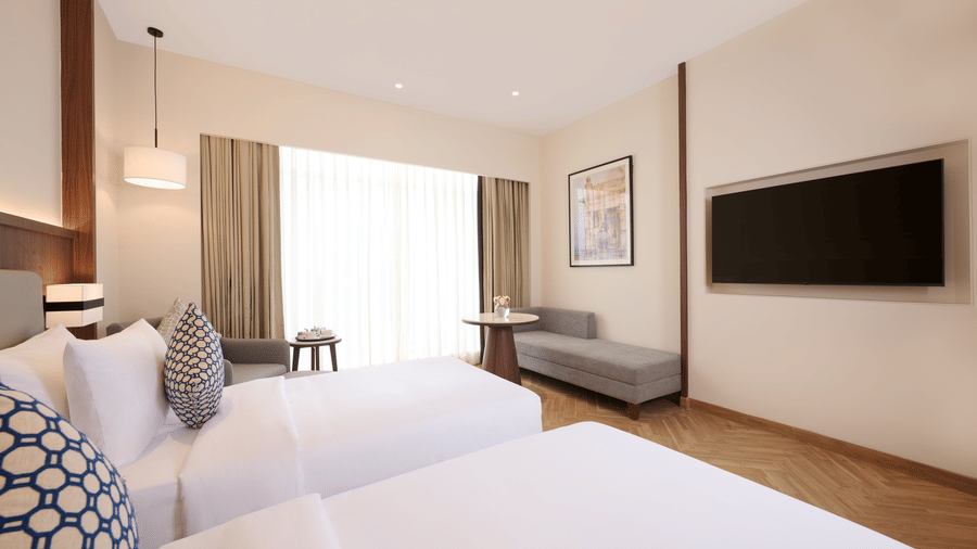 Skywings Sarovar Portico Mathura room wide view with two beds, patterned cushions, gray sofa, wall-mounted TV, and herringbone wood flooring.