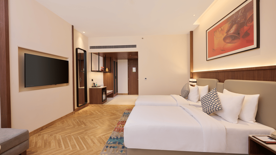 Skywings Sarovar Portico Mathura standard bed room entrance perspective with king bed, red wall art, herringbone wood flooring, and warm recessed lighting.