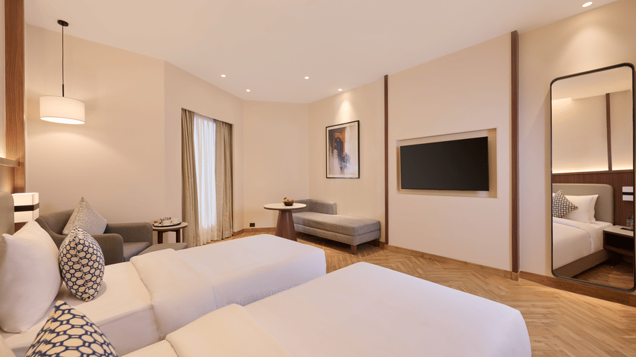Skywings Sarovar Portico Mathura standard twin room with two single beds, patterned cushions, wall-mounted TV, wooden flooring, and bright ambient lighting.