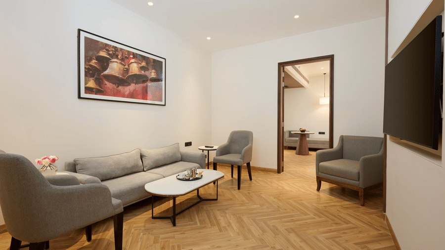 Skywings Sarovar Portico Mathura suite with king bed, gray armchairs, herringbone wood flooring, red framed wall art, and warm recessed lighting.