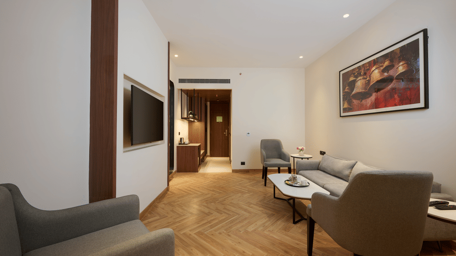 Skywings Sarovar Portico Mathura suite living area with sofa, work desk, wooden accent wall, framed art, herringbone flooring, and soft warm lighting.