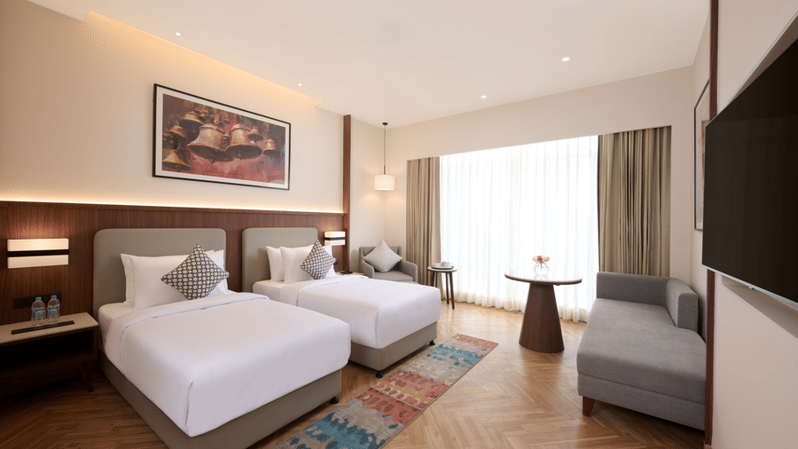 Skywings Sarovar Portico Mathura twin bed room with two single beds, gray sofa, patterned rug, wooden flooring, and warm ambient lighting.