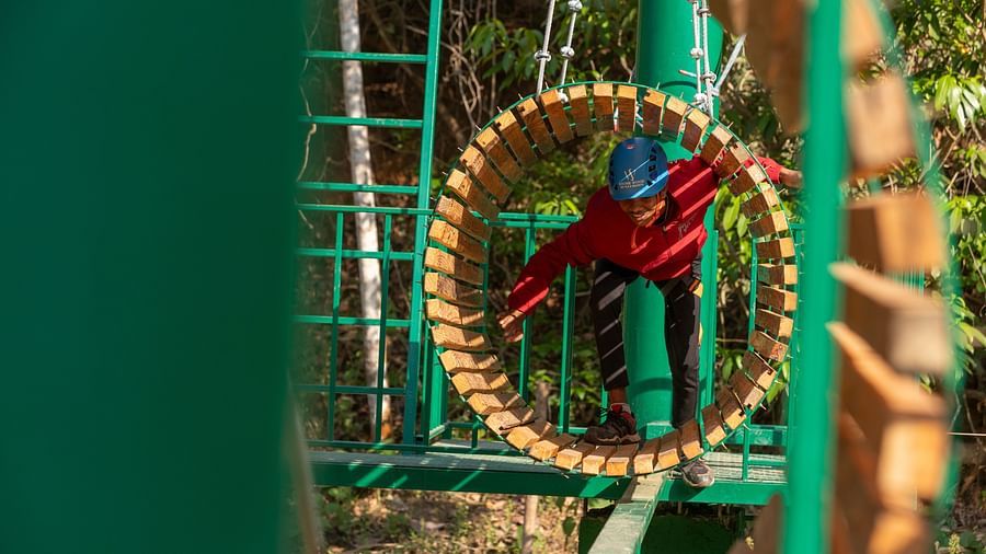Person wearing a blue helmet navigating through a circular wooden obstacle on a ropes course. | Stone Wood Jungle Resort, Dandeli