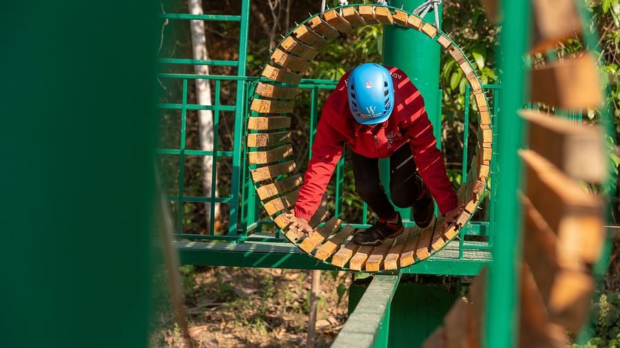 Person wearing a blue helmet navigating through a circular wooden obstacle on a ropes course. | Stone Wood Jungle Resort, Dandeli
